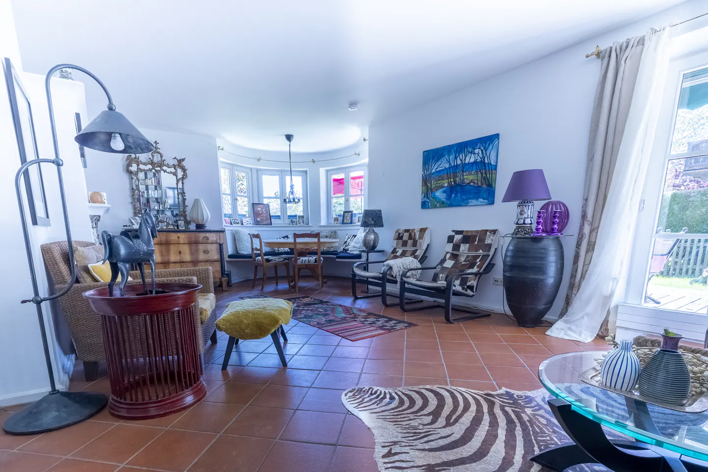 Bright living room with terracotta tile floors, zebra print rug, eclectic furniture, and a dining nook by bay windows.