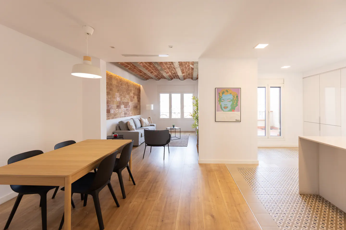 Bright, modern apartment interior with a wooden dining table, black chairs, and a gray sofa in the background. A brick wall and a Marilyn Monroe print add character.