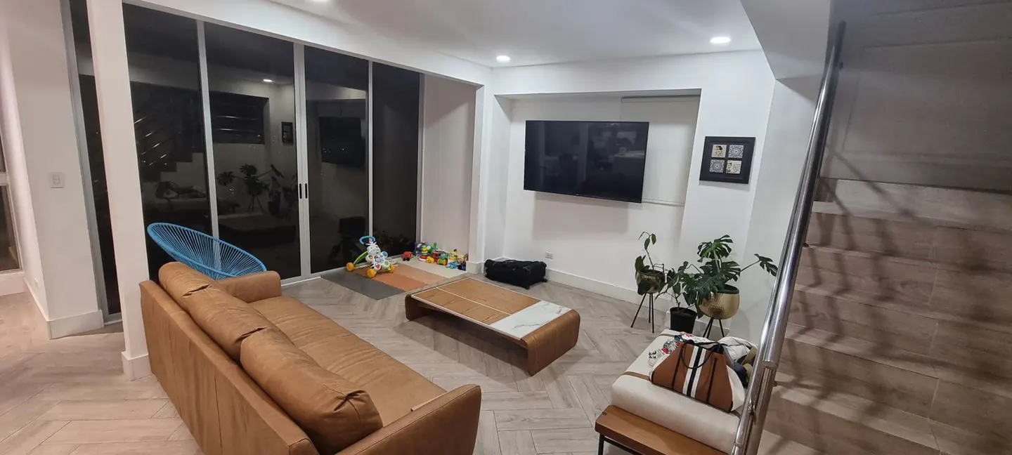 Modern living room with a tan leather sofa, coffee table, wall-mounted TV, and stairs, featuring light wood flooring.