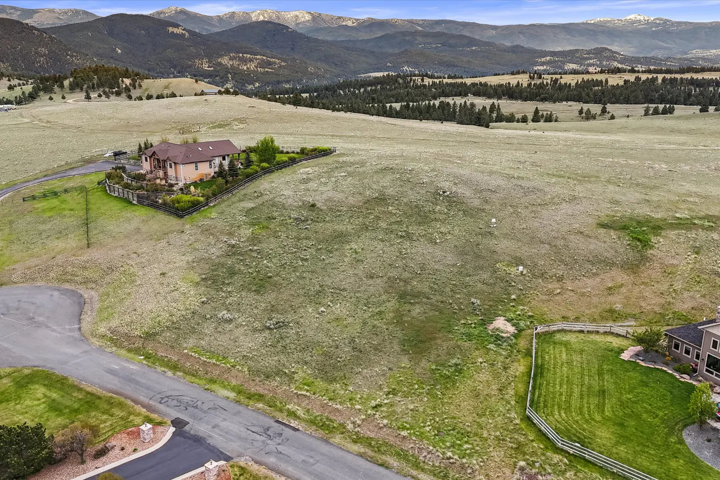 Aerial view of a tan house with a brown roof, surrounded by a wooden fence, in a grassy field with mountains in the background.
