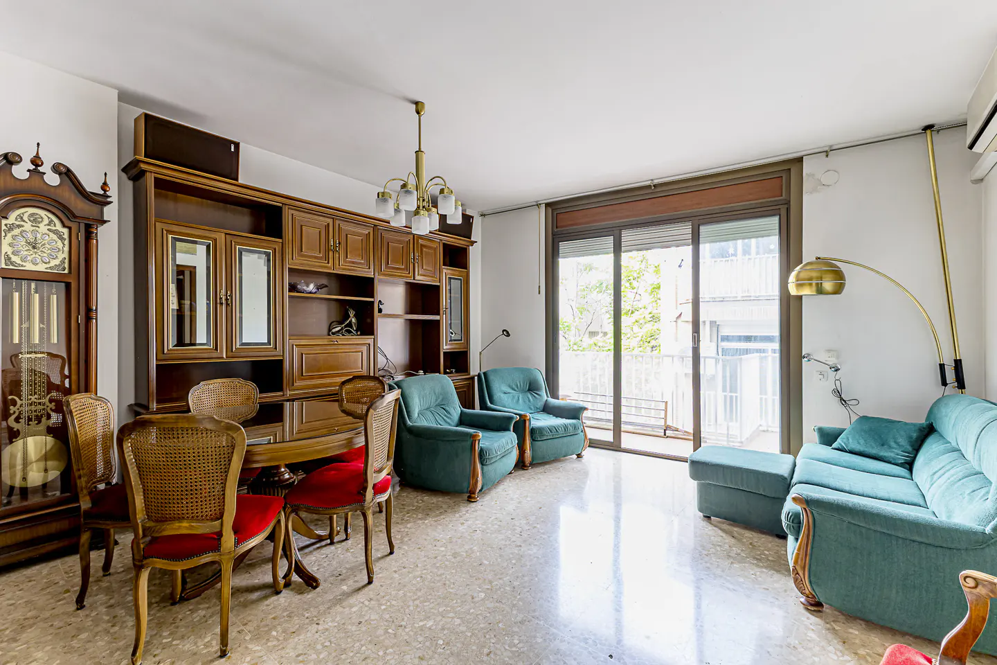 A living room with a grandfather clock, dining table, and teal sofa. A large wooden cabinet is against the wall.