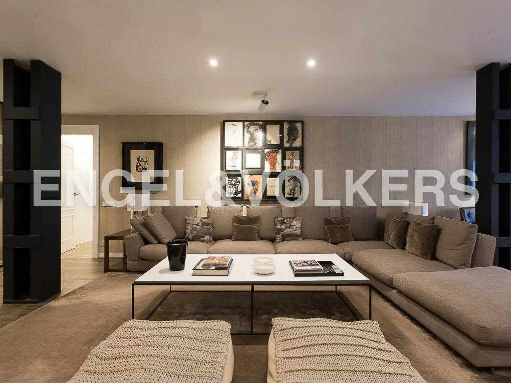 Living room with a large beige sectional sofa, white coffee table, and artwork on the wall. Two knitted poufs are in the foreground.