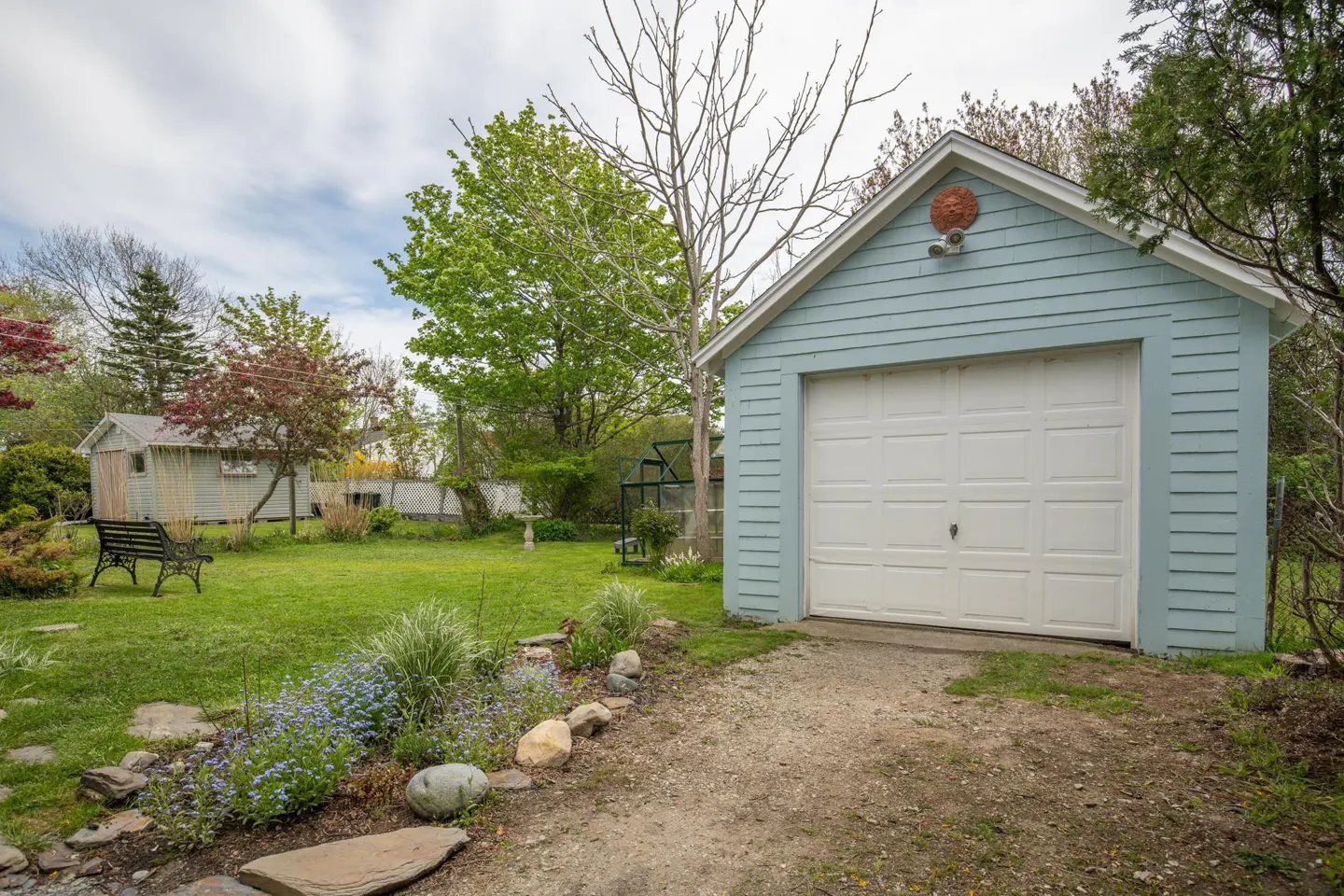 A light blue garage with a white door sits on a gravel driveway, surrounded by a green lawn and trees.