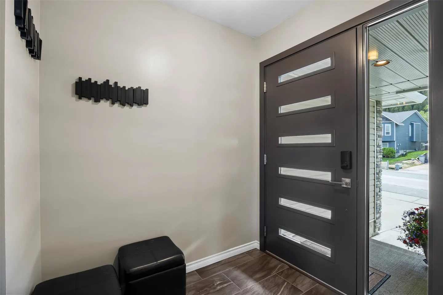 Entryway with a modern gray door with horizontal windows, black coat rack, and black ottomans on a wood-look tile floor.