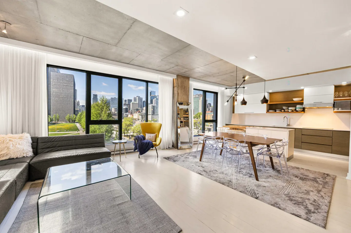 Bright, open-concept condo with a gray sectional, glass coffee table, yellow chair, and dining table with clear chairs. Large windows offer city views.