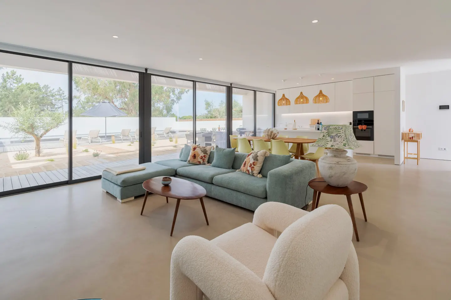Bright, open-plan living room with a large blue sectional sofa, white armchair, and floor-to-ceiling windows overlooking an outdoor patio.