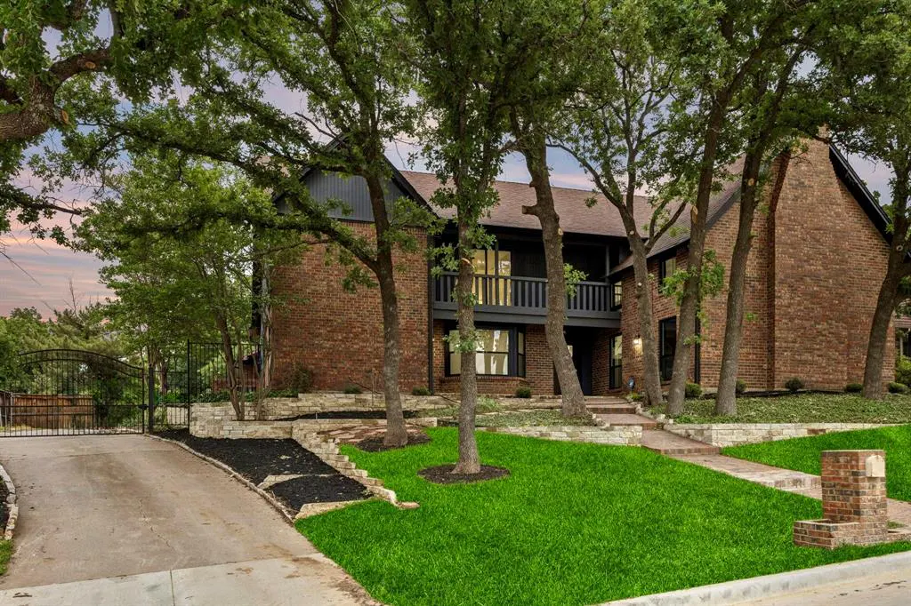 Two-story brick house with black trim, a balcony, and a gated driveway, surrounded by green trees and grass.