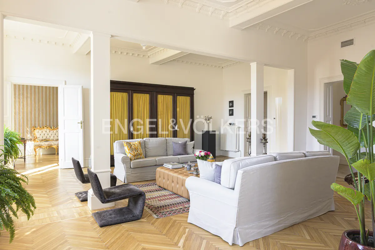 Bright living room with hardwood floors, white walls, and tall ceilings. Two sofas face each other over a brown ottoman on a patterned rug. Two gray chairs are in the foreground.