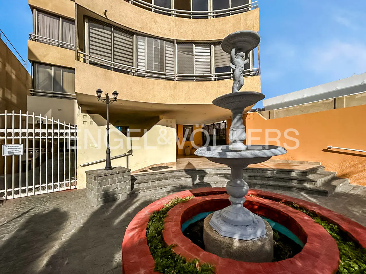Exterior view of a tan apartment building with a fountain in the foreground. The fountain has a cherub statue on top. A white gate is to the left.
