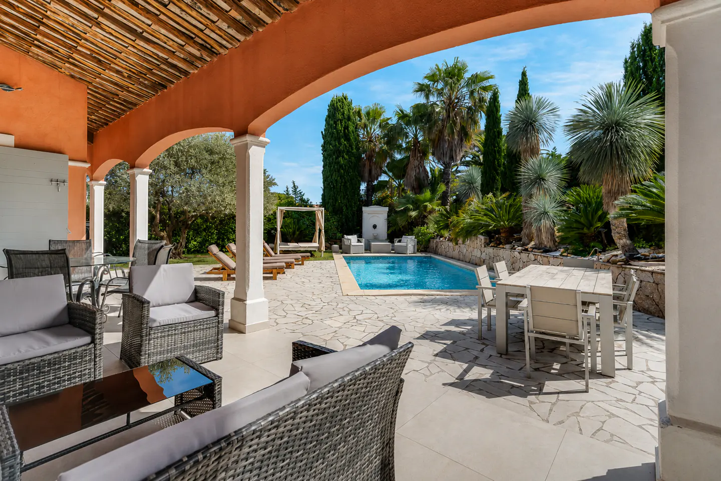 Covered patio with wicker furniture overlooking a sunny pool area with palm trees and a stone-paved terrace.