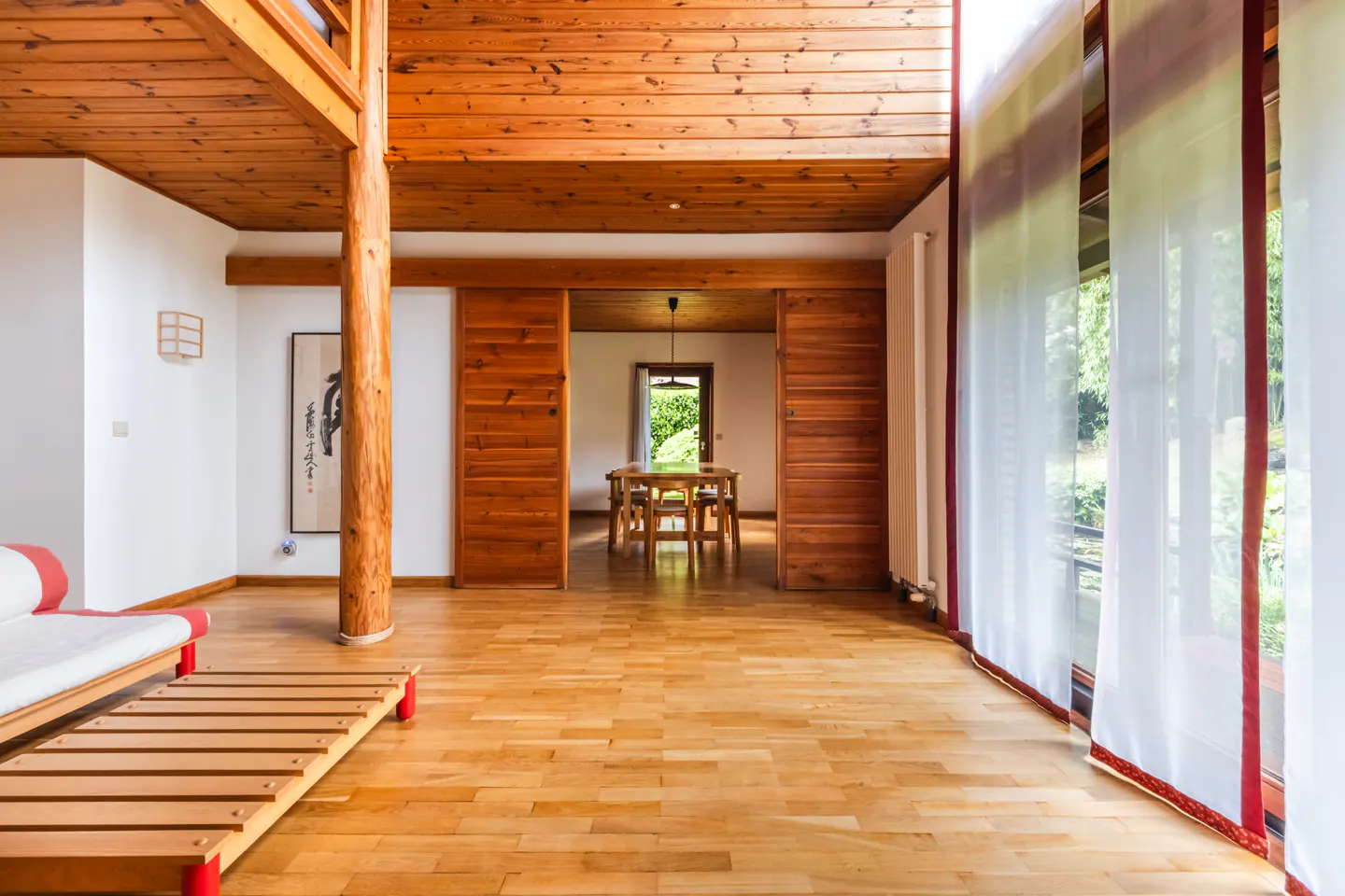 Bright, open living space with wood floors, ceiling, and sliding doors. White walls, sheer curtains, and a glimpse of a dining room beyond.