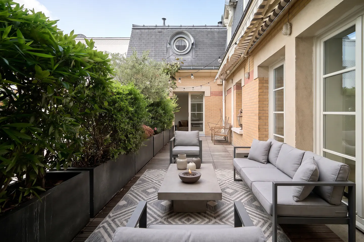 Outdoor patio with gray furniture, a patterned rug, and green plants in black planters. A building with windows and a round window is in the background.