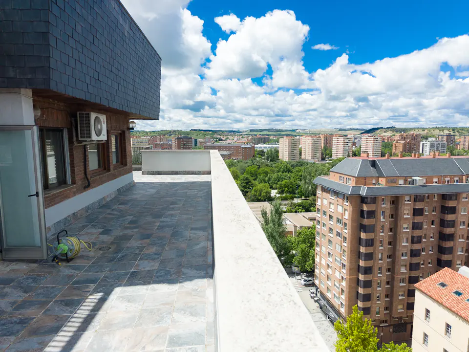Rooftop view of a city with blue sky and clouds. Tiled floor, white railing, and brick building with windows are visible.