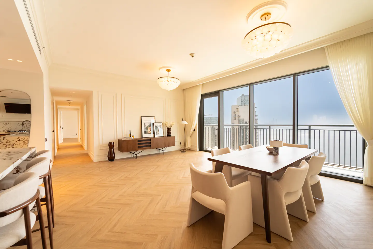 Bright, modern apartment interior with wood floors, dining table, and city views from large windows. Cream walls and elegant lighting.