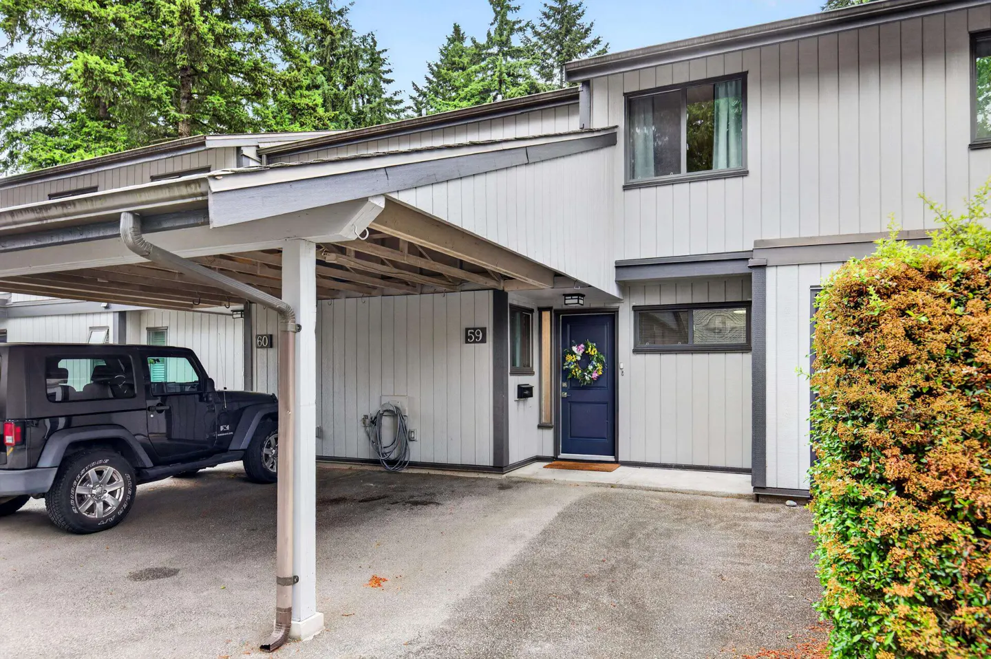 Exterior of a grey townhouse with a carport, a black Jeep parked underneath, and a blue front door with a floral wreath.