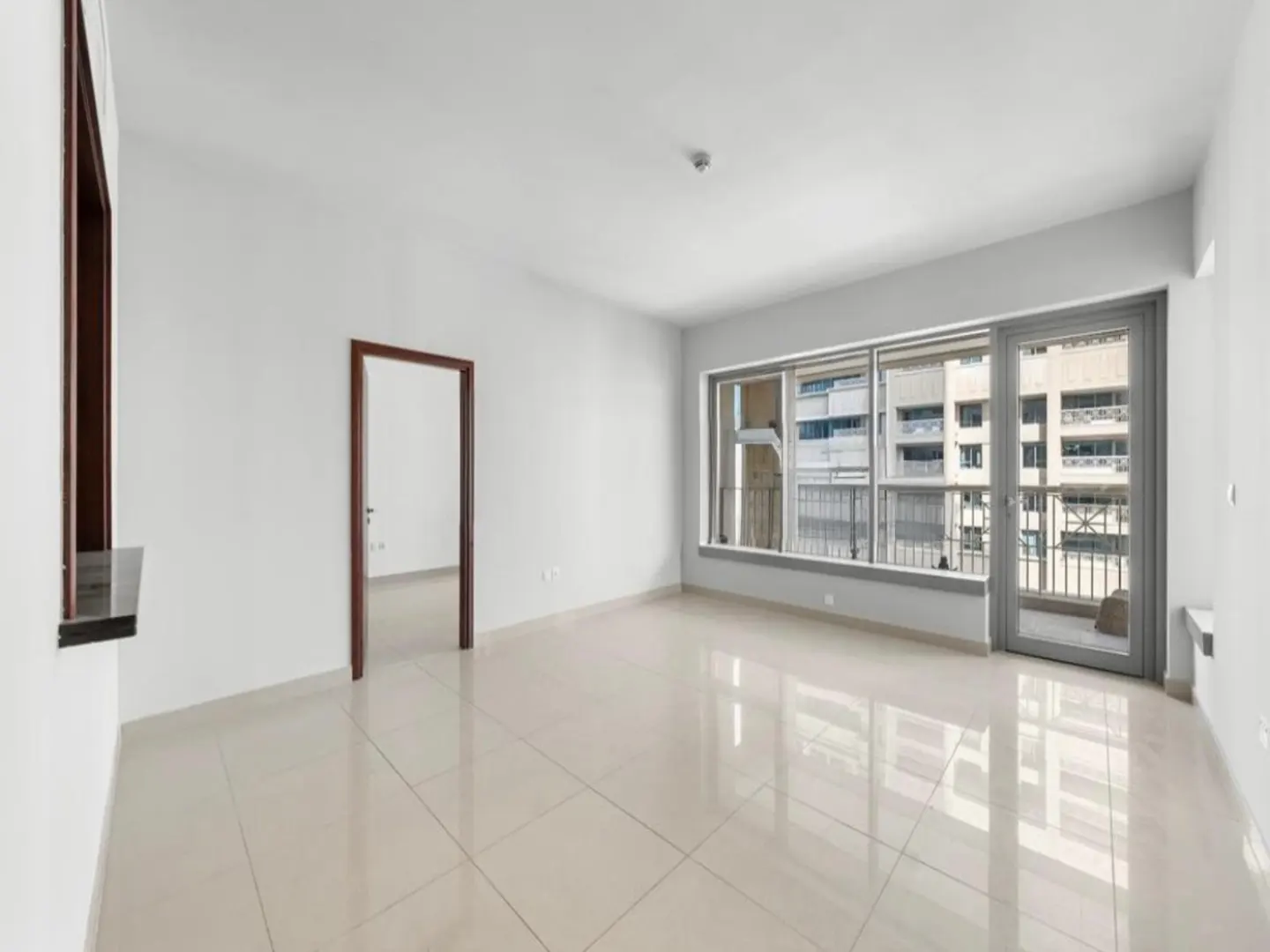 Bright, empty room with white walls, tile floor, and brown door frames. A large window overlooks a city building.