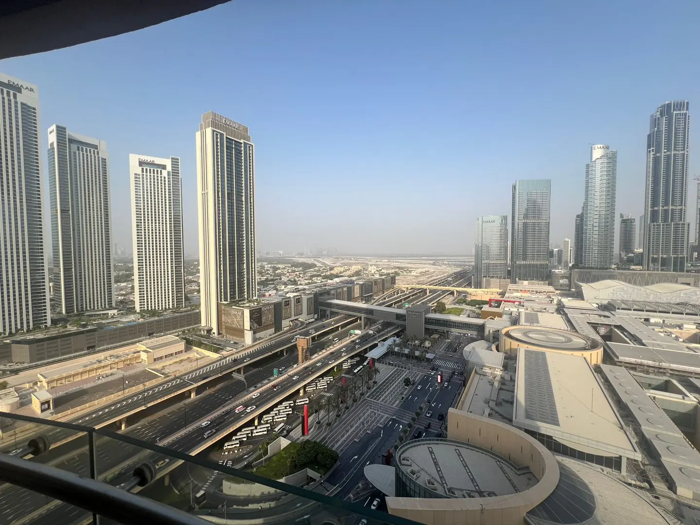 Cityscape view from a high-rise balcony. Tall buildings, highways, and streets stretch into the distance under a clear blue sky.