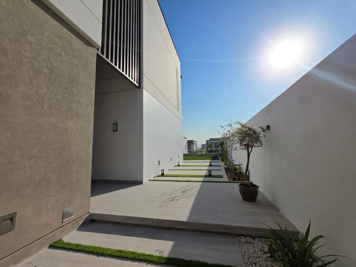 Modern home exterior with a walkway lined with grass, trees, and white walls under a bright blue sky.