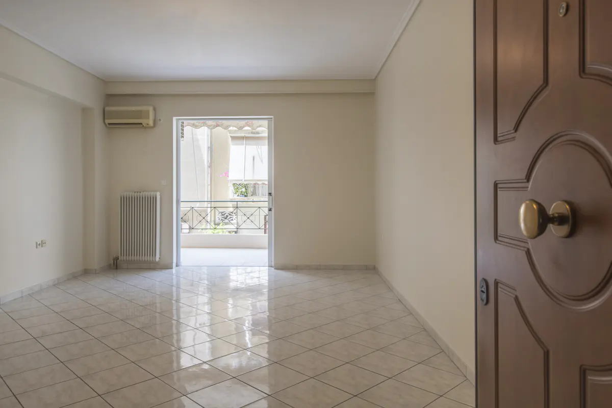 Empty room with beige walls, tiled floor, radiator, air conditioner, and a balcony door, viewed from an open wooden door.