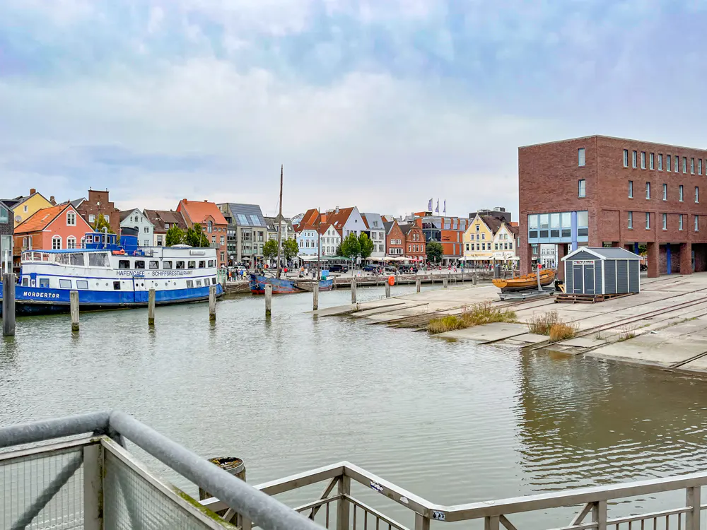 A harbor scene with a blue and white ferry, colorful historic buildings, and a modern brick building under a cloudy sky.