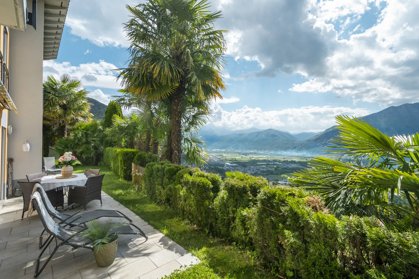 Outdoor patio with lounge chairs, table, and garden overlooking a valley with mountains in the background under a blue sky with white clouds.