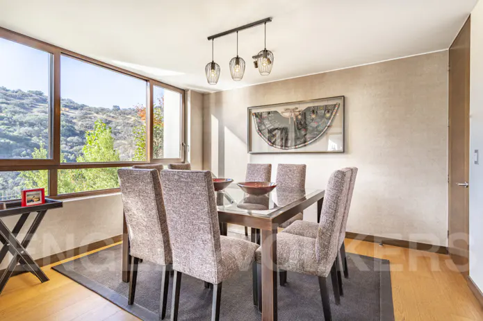 Dining room with a glass table, six upholstered chairs, and a view of green hills through a large window.