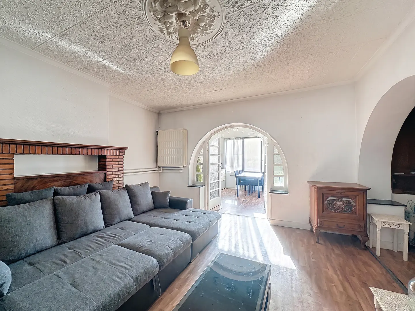 Living room with a gray sectional sofa, brick fireplace, and arched doorway leading to a dining area with a table and chairs.