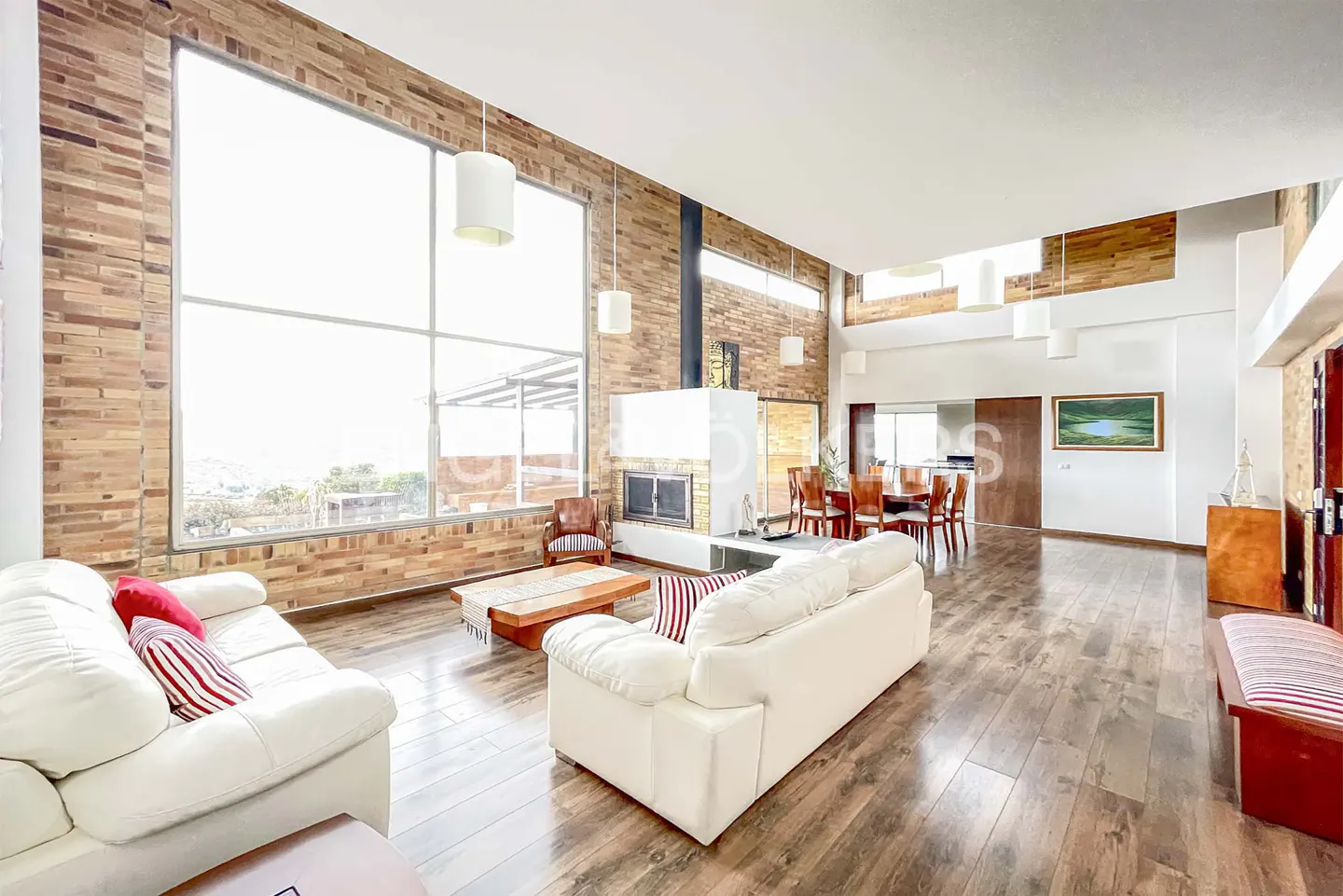 Bright living room with brick walls, hardwood floors, and white sofas. Large window with a view. Fireplace and dining area visible.