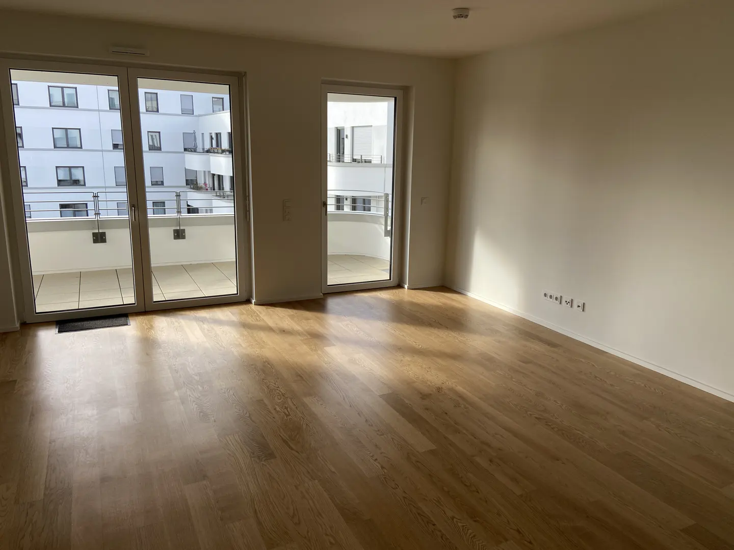 Bright, empty room with light wood floors and white walls. Two sets of glass doors lead to balconies. Building visible outside.