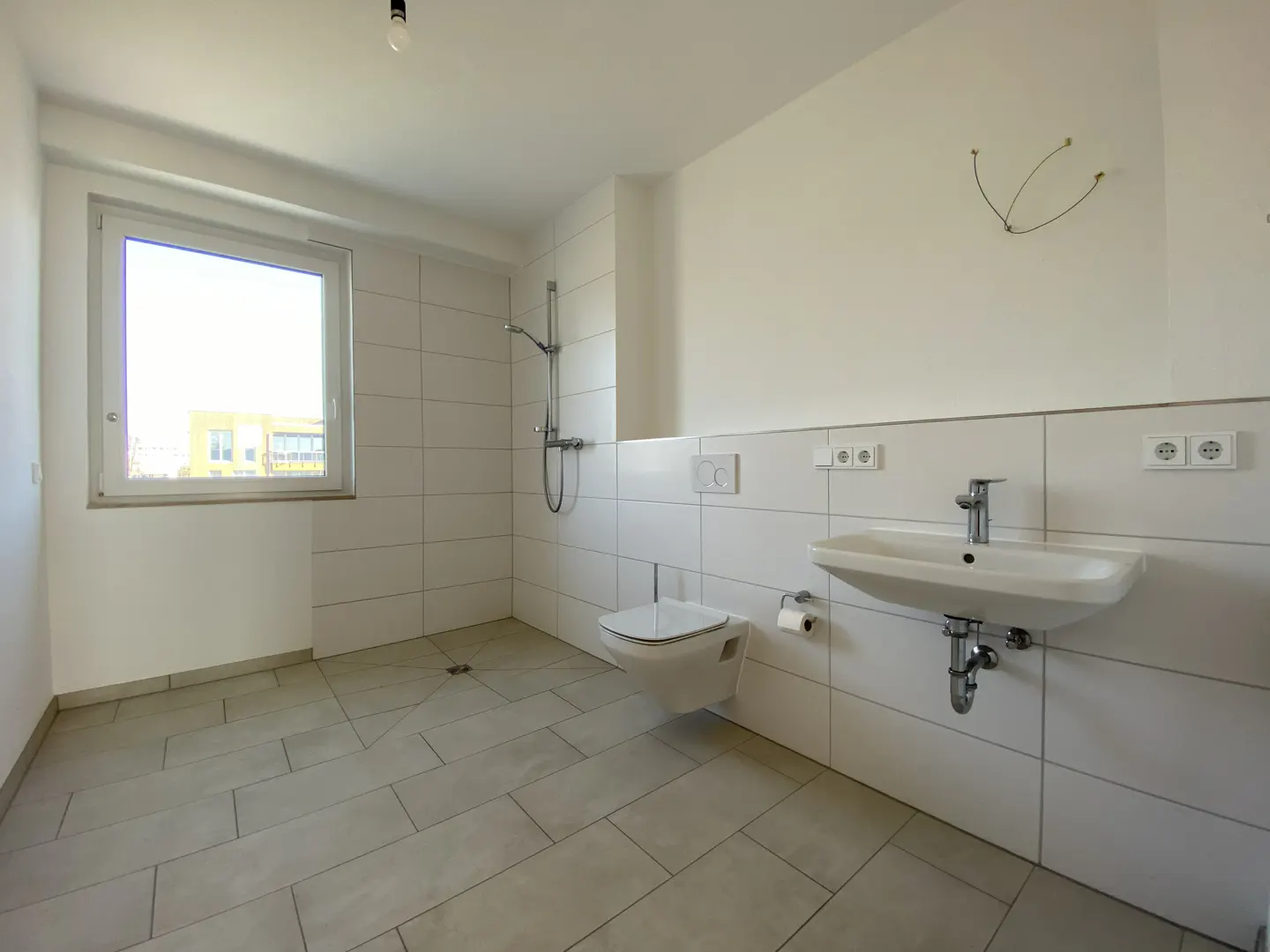 Bright, modern bathroom with white tiled walls, gray tiled floor, toilet, sink, and shower. A window lets in natural light.
