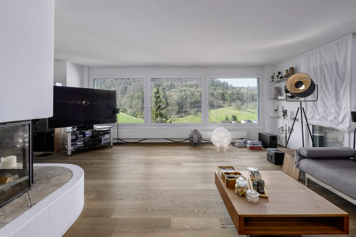 Bright living room with wood floors, a fireplace, and a large TV. Three windows offer a view of green hills. A wooden coffee table sits in front of a gray sofa.