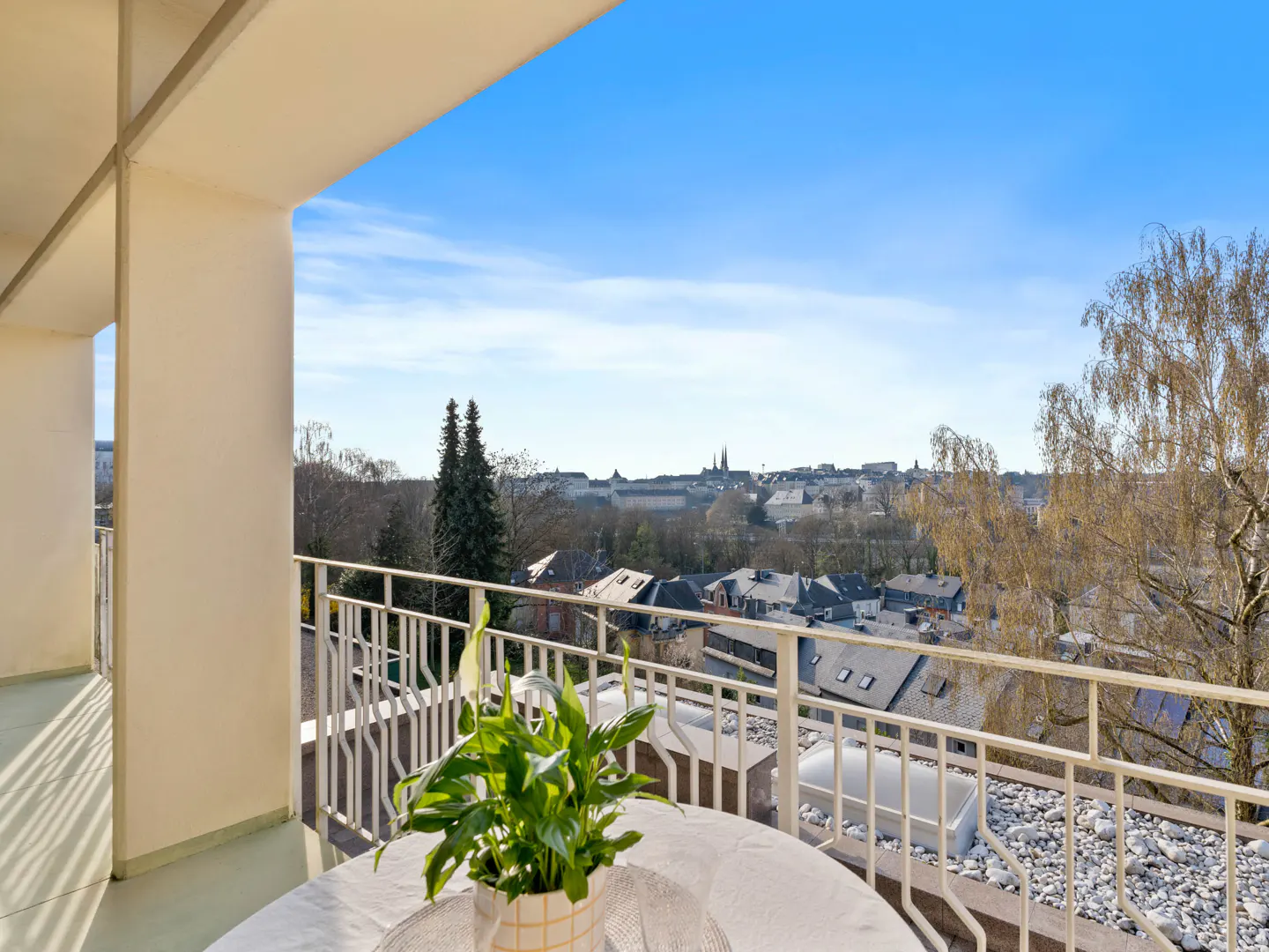 View from a balcony with a white table and potted plant, overlooking a town with trees and a clear blue sky.