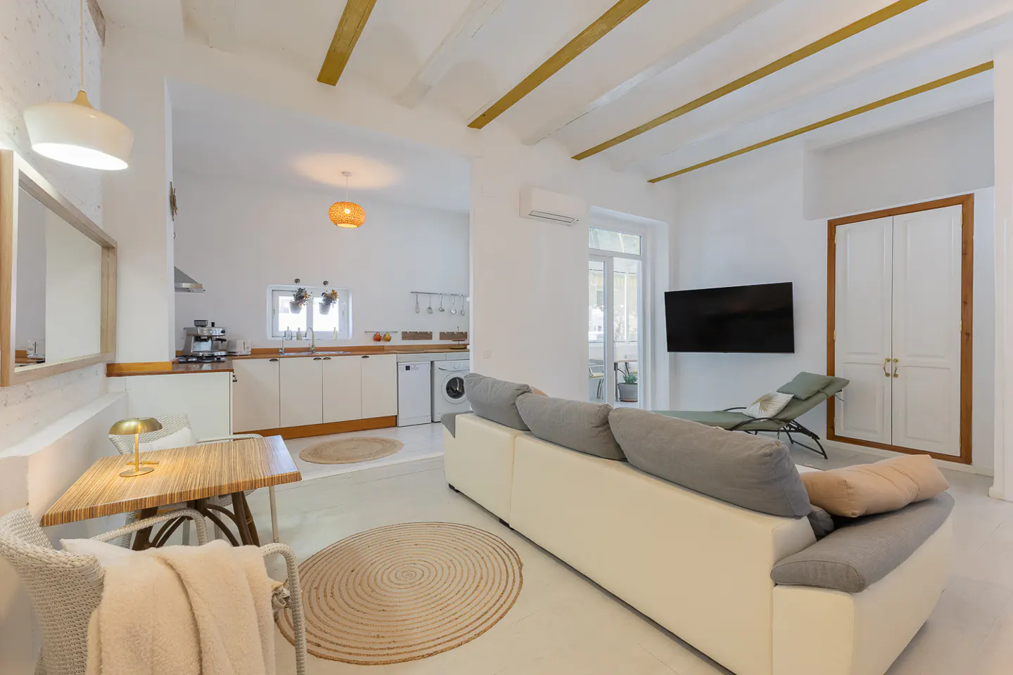 Bright, open-concept living space with white walls, wood beams, and a modern kitchen. A cream sofa faces a TV, with a lounge chair nearby.