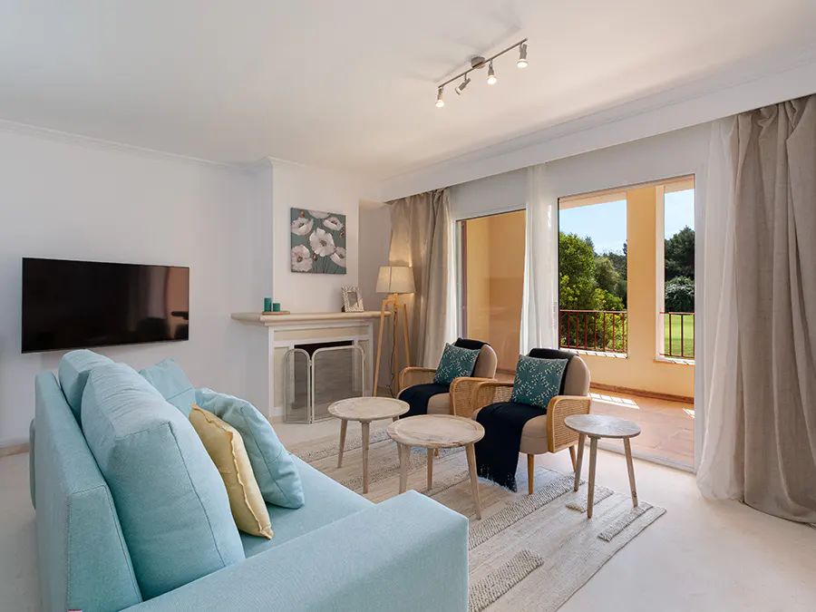 Bright living room with a light blue sofa, two chairs, and a balcony view of trees. A TV hangs on the wall.