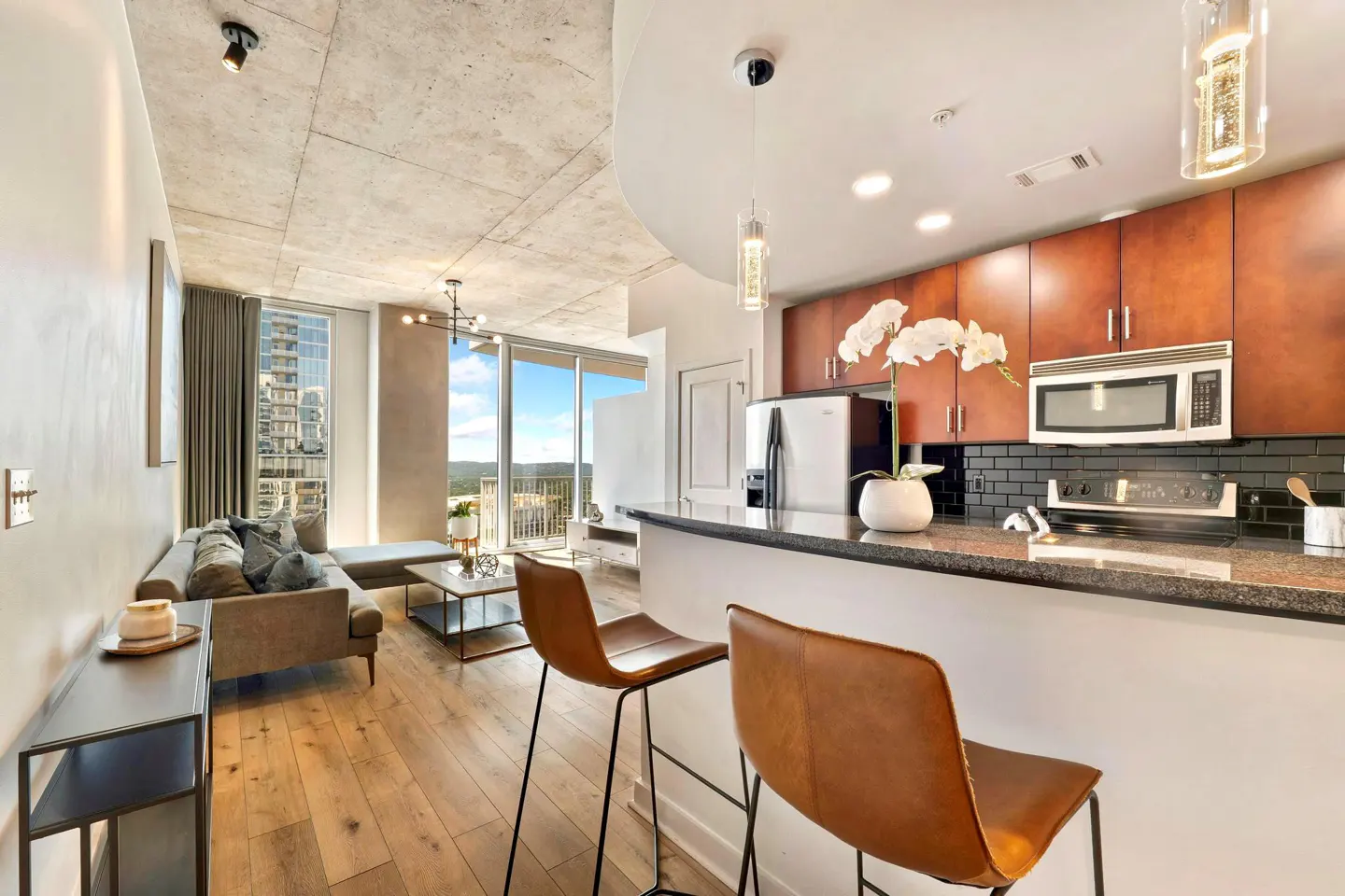 Modern open-concept apartment with a kitchen featuring brown cabinets, a breakfast bar, and a living area with city views.