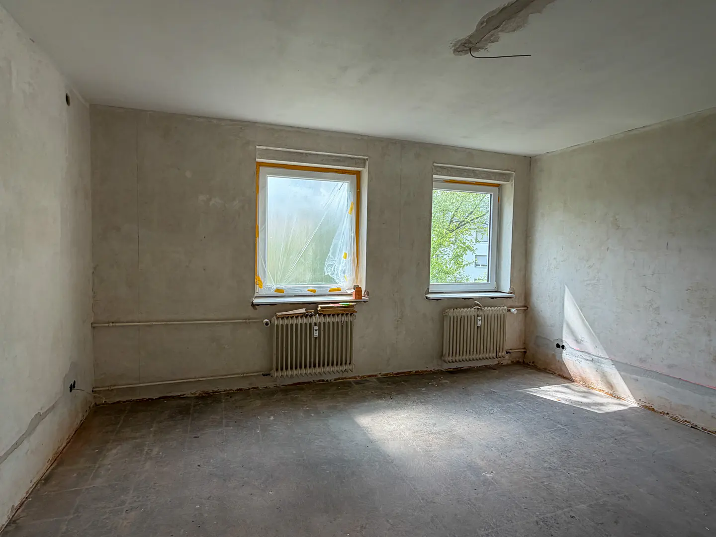 Unfinished room with two windows, one covered in plastic, and radiators below. Walls are plaster, floor is concrete.