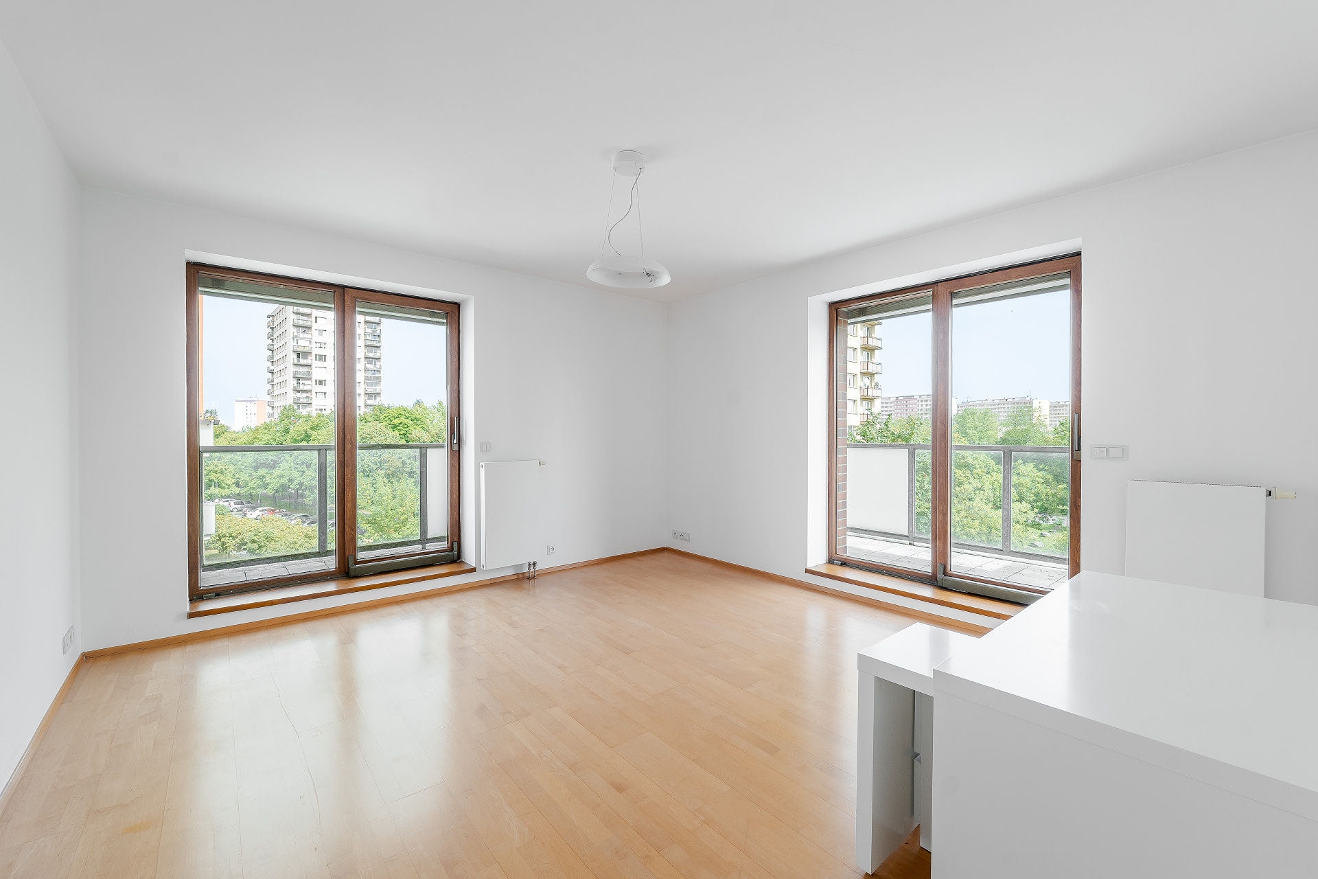 Bright, empty room with light wood floors, white walls, and two large glass doors leading to balconies with city views. [AI-generated text]