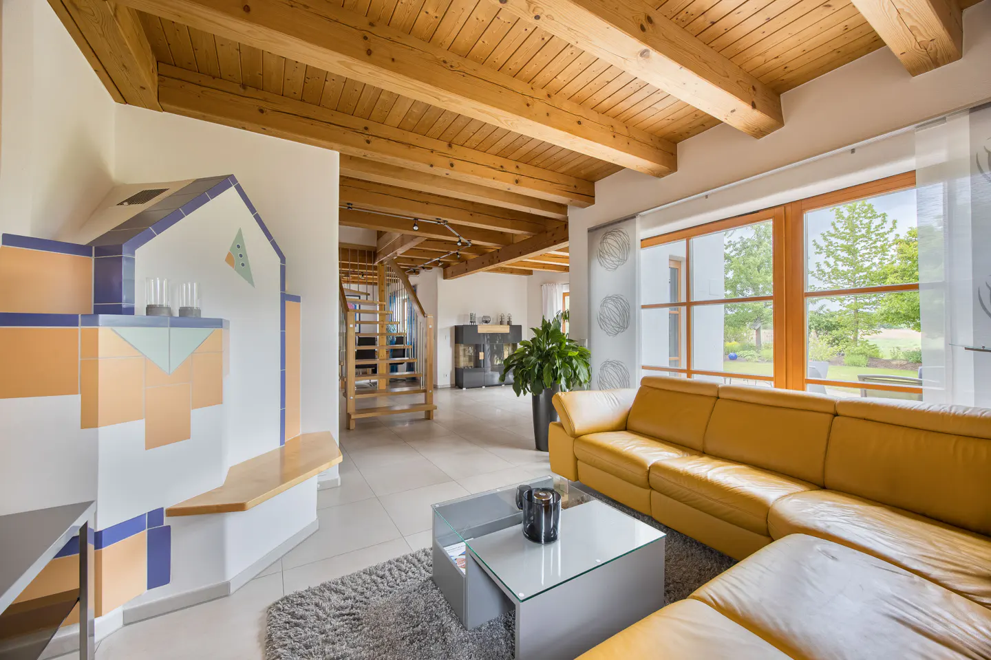 Bright living room with wood-beamed ceiling, yellow leather sofa, glass table, and tiled wall feature. Staircase visible in background.