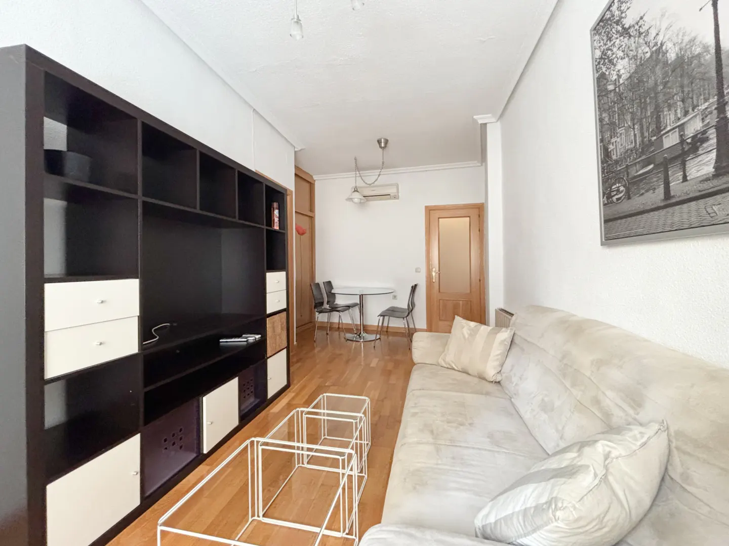 Living room with a large black shelving unit, beige sofa, wire coffee tables, and a dining area with a table and chairs.