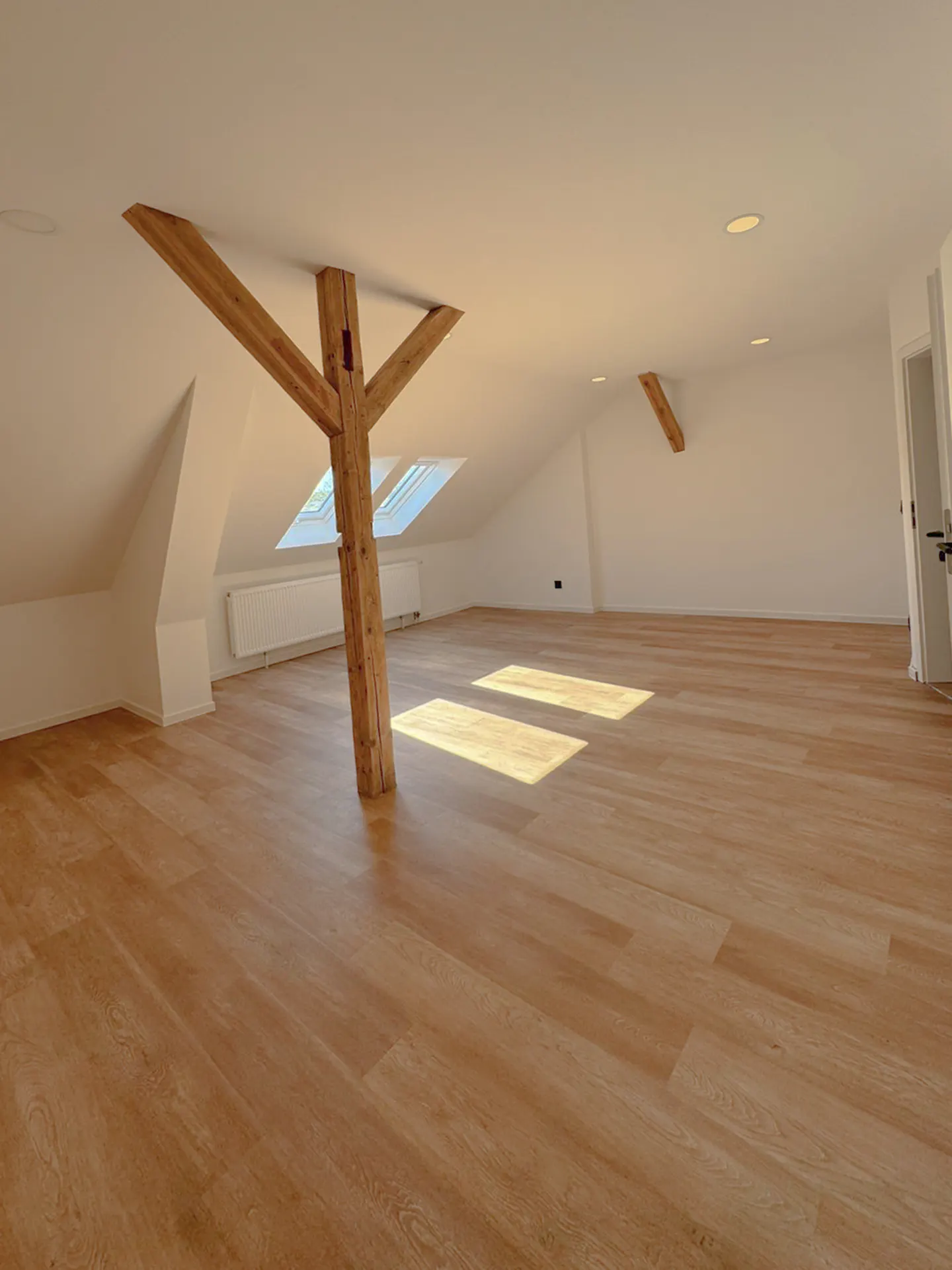 Attic room with light wood floors, white walls, and exposed wooden beams. Sunlight streams in from skylights.