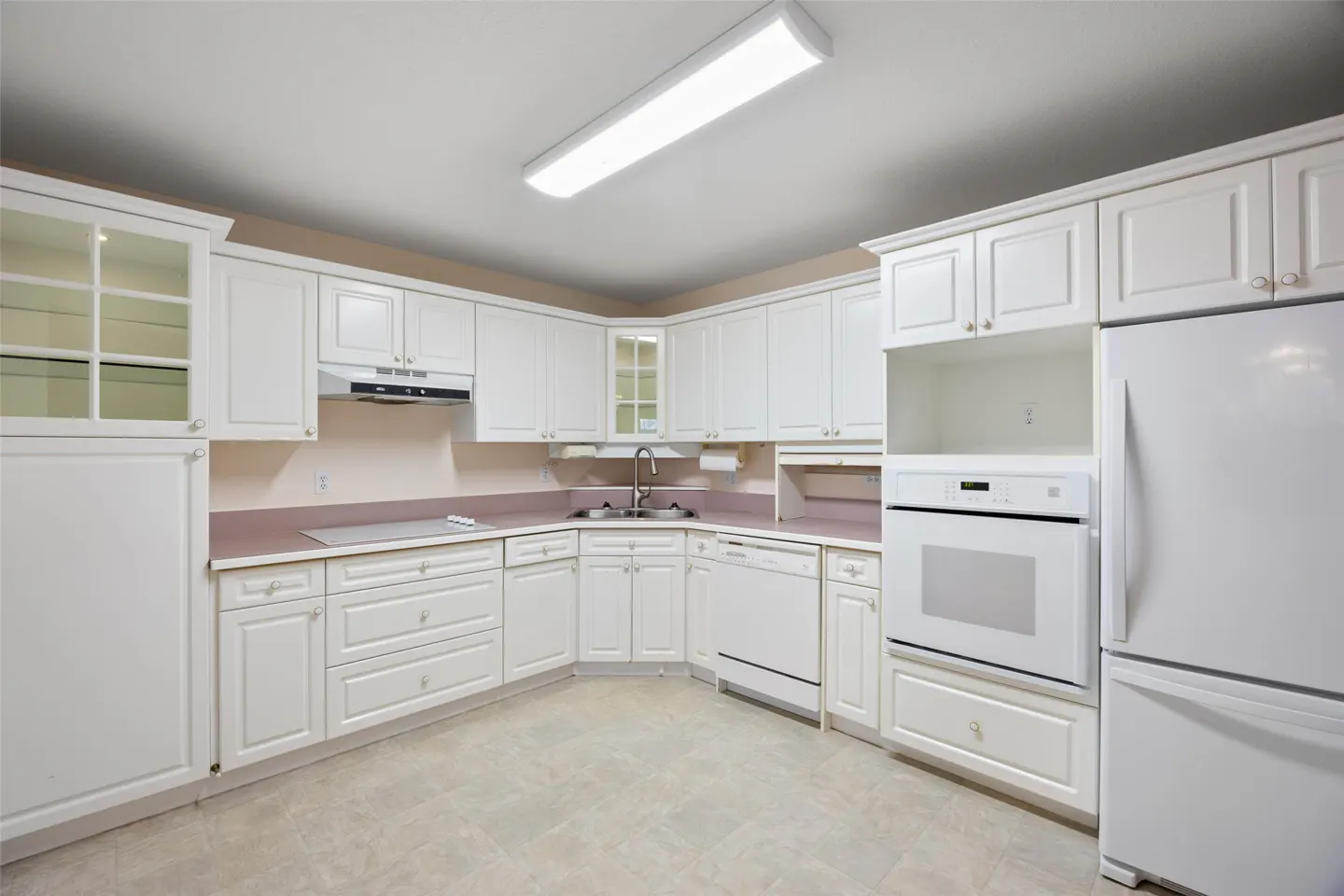 Bright kitchen with white cabinets, appliances, and light pink countertops. A fluorescent light illuminates the room.