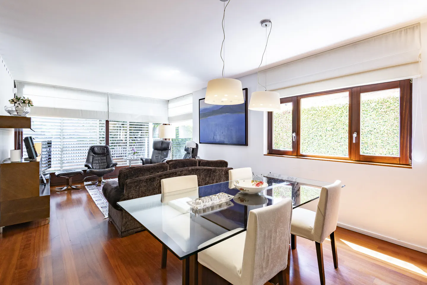 Bright living room with hardwood floors, a glass dining table with white chairs, and a brown sofa. Large windows offer a view of greenery.