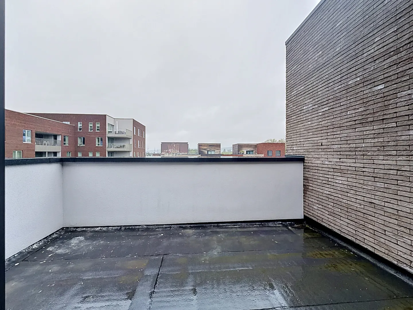 Empty rooftop terrace with a white wall, brick side wall, and a view of neighboring apartment buildings under a gray sky.
