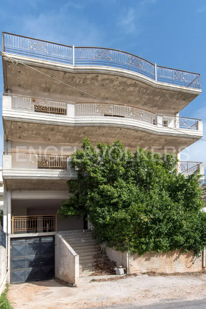 Three-story concrete building with large balconies, metal railings, a ground-level garage, and a large green tree.
