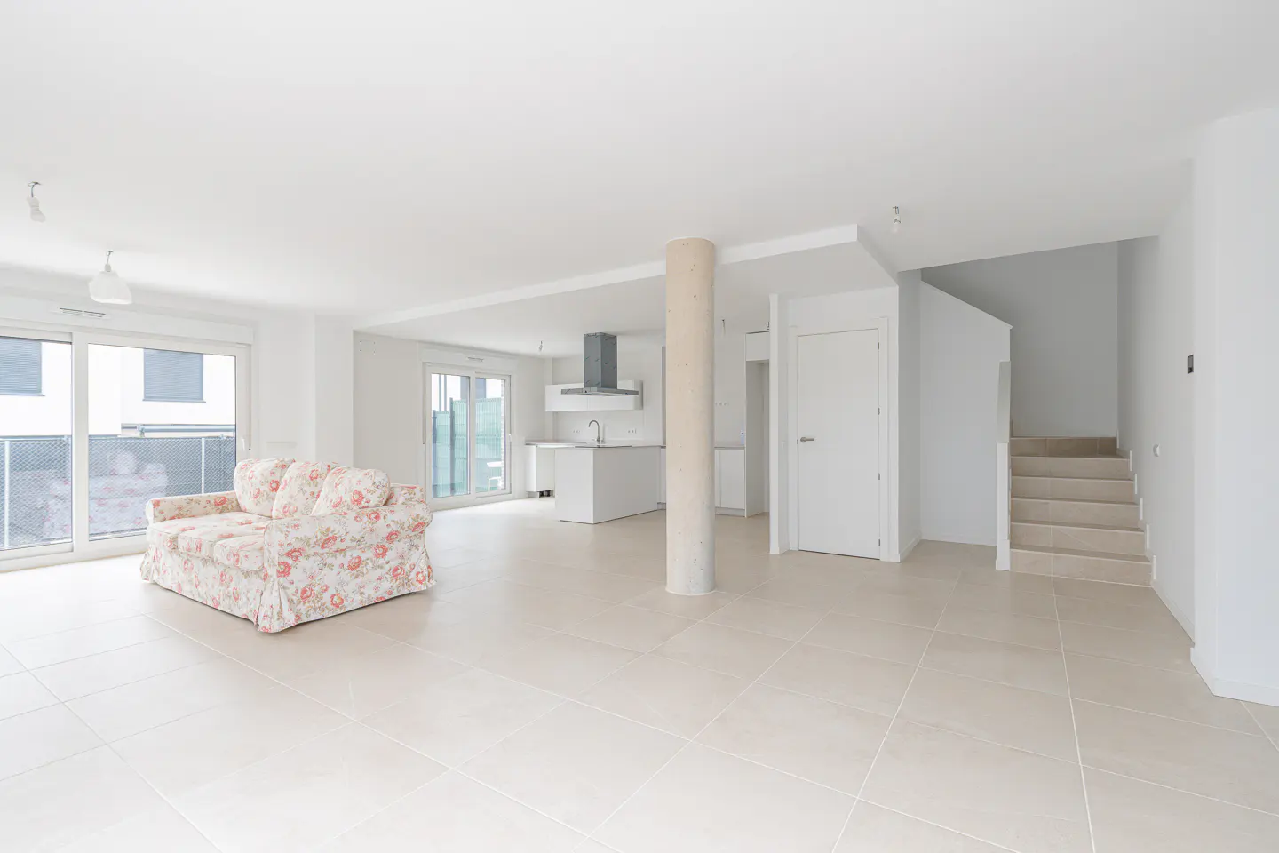 Bright, open-plan living space with floral sofa, kitchen island, and stairs. White walls and tile floors create a modern, airy feel.