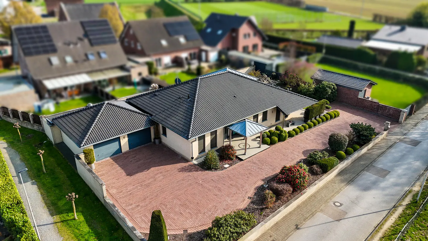 Aerial view of a beige house with a dark gray roof, blue garage doors, and a brick driveway. Green bushes line the driveway. Other houses are in the background.