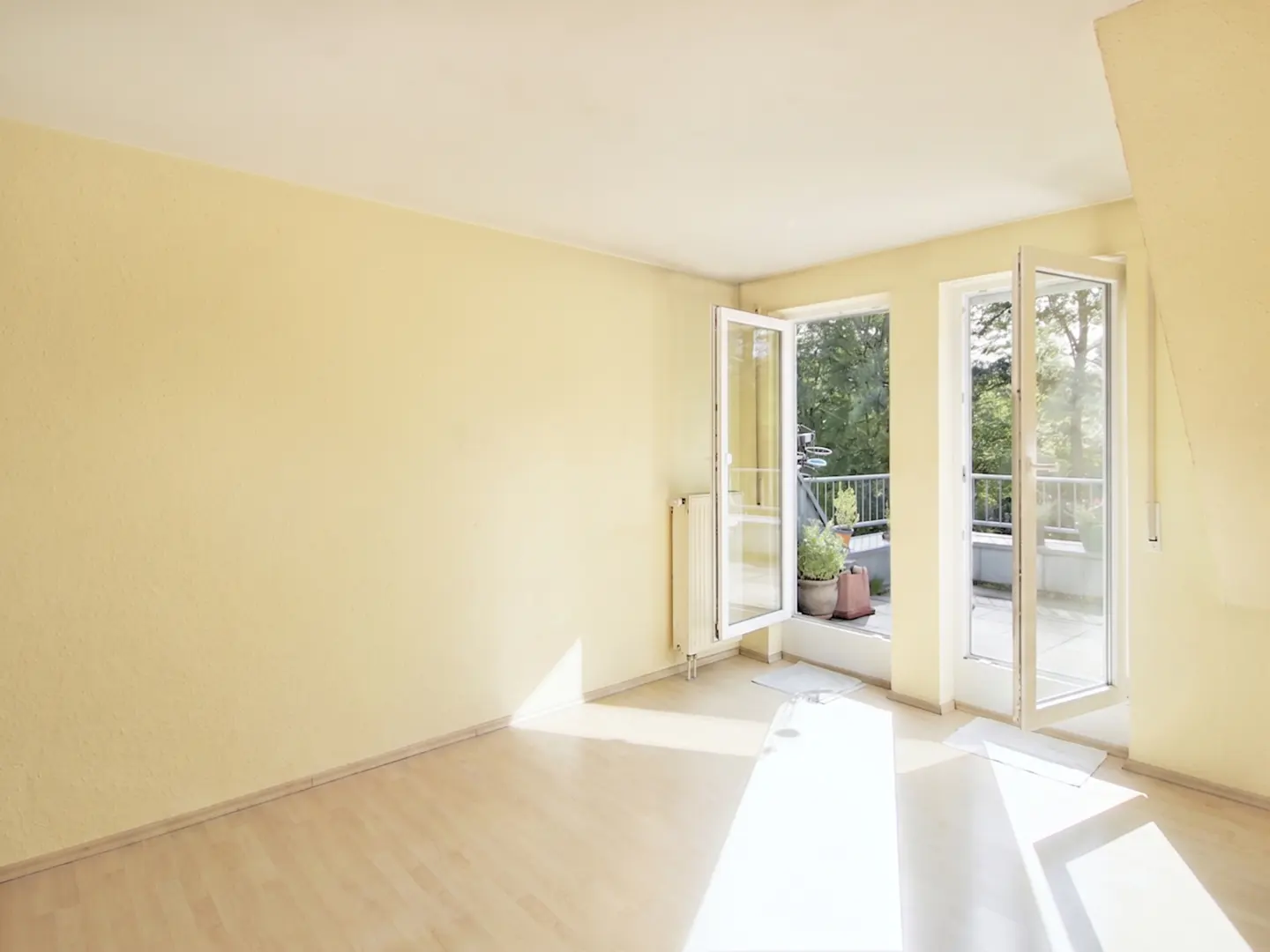 Bright, empty room with light yellow walls and wood floor. Open glass doors lead to a balcony with plants and trees visible outside.