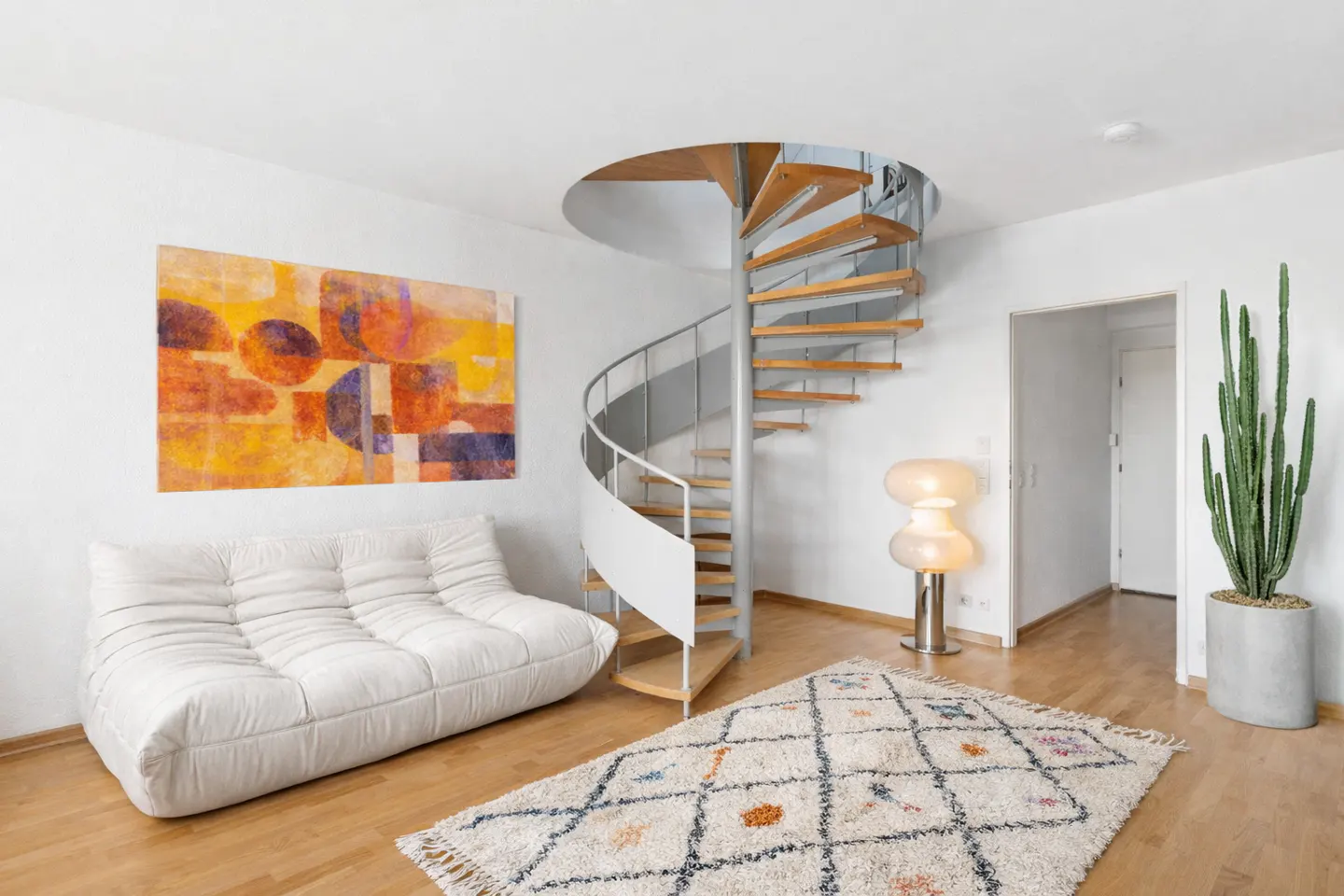 Bright living room with a white sofa, colorful art, and a spiral staircase. A patterned rug and tall cactus add texture.