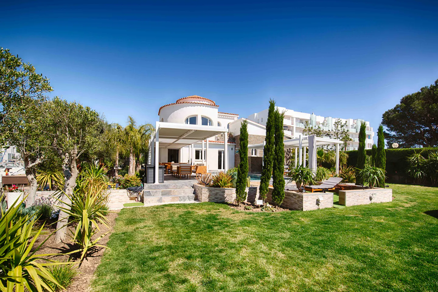 A white house with a red-tiled dome roof, a covered patio with a table and chairs, and a green lawn.
