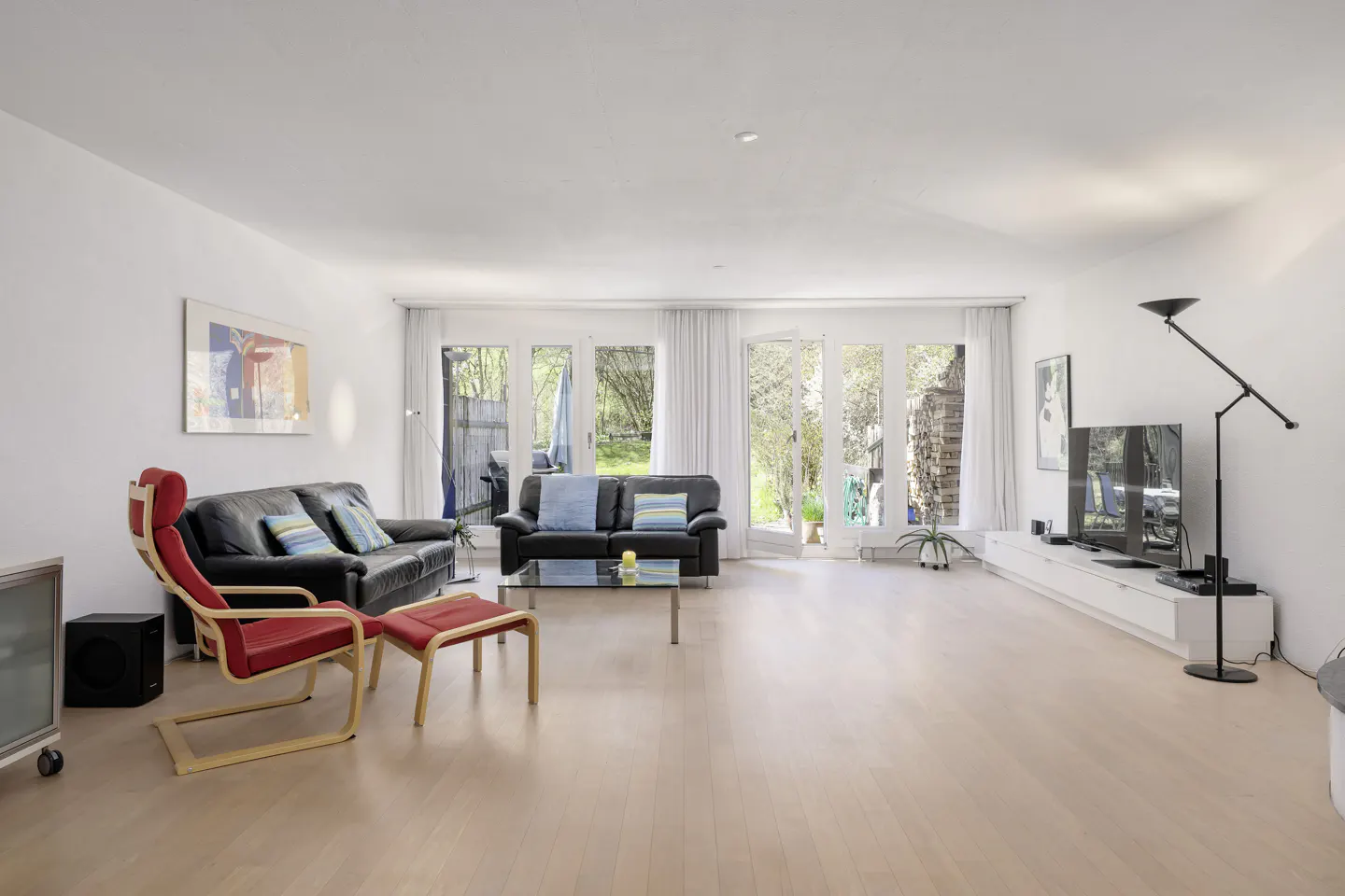 Bright living room with light wood floors, white walls, and large windows. Black leather sofas, red chair, TV, and modern decor.